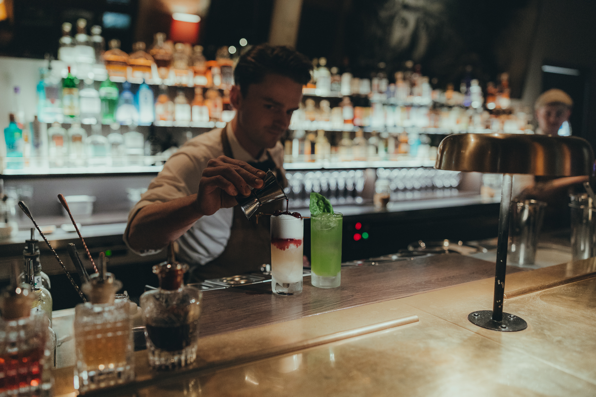 A bartender with two highballs: a Gin Basil Smash and a New York Sour highball. He is adding the wine on top of the foam of the New York Sour highball.