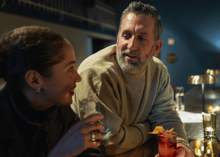 Ein Mann und eine Frau sitzen an einer schwach beleuchteten Bar und lächeln sich warm an. Er hält einen Cocktail mit einer Orangengarnitur in der Hand, während sie ein leichtes Getränk trinkt. Intimes und gemütliches Ambiente.